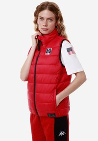 Gilet imbottito rosso con colletto alto, chiusura frontale con zip e due tasche laterali. Presenta una patch "US" e una piccola bandiera americana sulla manica.