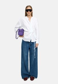 Biała koszula z detalem w postaci zebranej wiązania, szerokie niebieskie dżinsy, fioletowa torebka i czarne oversized okulary przeciwsłoneczne, noszone z burgundowymi butami.