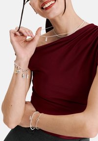 Silberschmuck präsentiert zu einem burgunderfarbenen Oberteil. Auffällige Details sind geschichtete Halsketten, ein Charms-Armband und elegante Texturen.