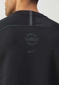 Černé sportovní tričko s kulatým výstřihem, s tonálním logem Nike a grafikou "STRIVE FOR GREATNESS" na zádech. Hladký povrch.
