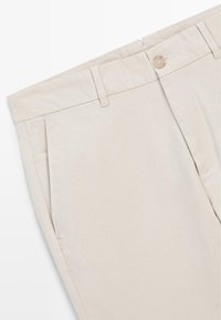 Pantalon beige avec poches avant, passants de ceinture et fermeture à bouton, posé à plat, montrant la partie supérieure de la taille et les hanches.