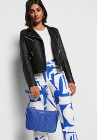 Giacca cropped in pelle nera, camicia bianca, pantaloni blu e bianchi a fantasia e una borsa blu trapuntata con tracolla.