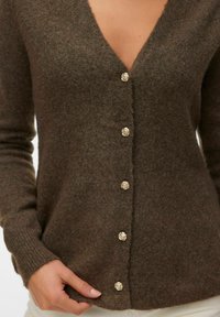 Brauner Strickcardigan mit fünf goldenen, blumenförmigen Knöpfen, getragen von einer Person, die mit einer Hand den Saum richtet.