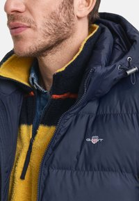 Marineblaue Steppjacke mit gestepptem Muster, kombiniert mit einer gelb-schwarzen Fleece-Innenschicht, die einen Reißverschluss und ein Logo-Detail aufweist.