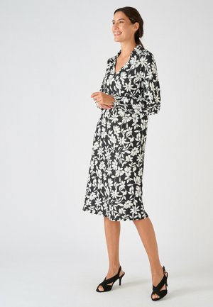 Donna sorridente, indossa un vestito midi nero con stampa floreale bianca e maniche lunghe, abbinato a tacchi neri open-toe, in piedi di profilo.