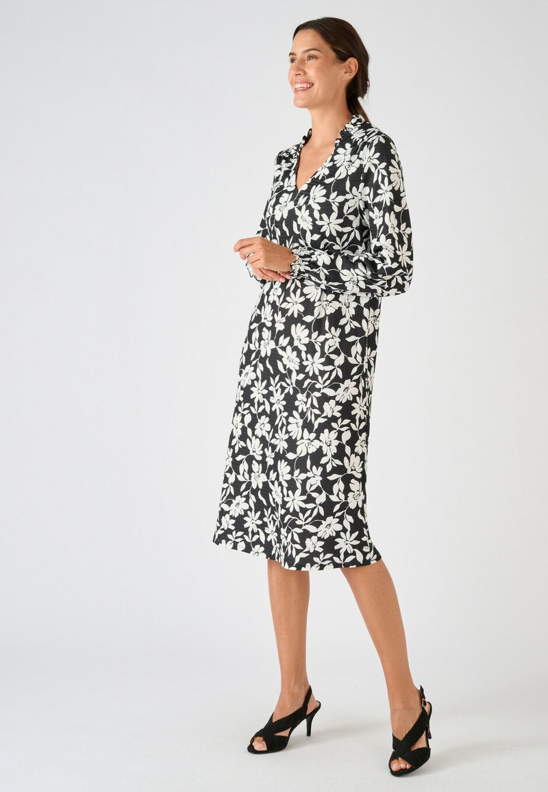 Femme souriante, portant une robe midi noire à imprimé floral blanc et manches longues, associée à des talons ouverts noirs, debout de profil.