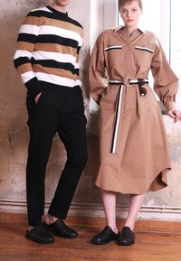 Calças cargo pretas e uma camisa de manga longa riscada castanha em uma pessoa; um vestido camisa bege com cinto na outra, ambos combinados com tamancos pretos.