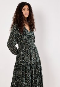 Woman with long curly hair wearing a dark patterned long-sleeve dress with a gathered waist and V-neck, standing against a plain background.