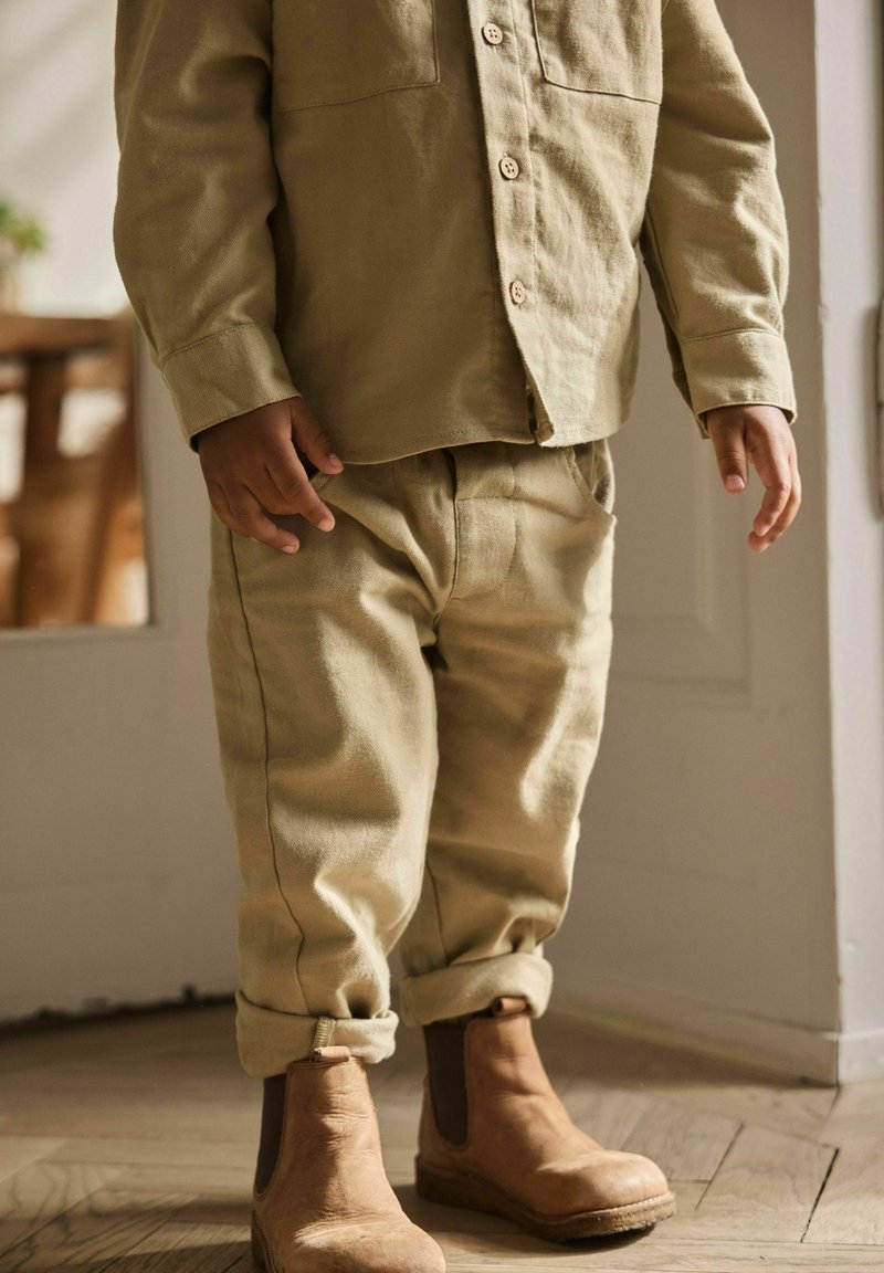 Enfant portant un pantalon beige, une chemise boutonnée assortie et des bottines marron, debout à l'intérieur sur un sol en bois près d'un mur blanc.