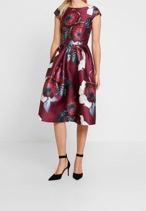 Woman wearing a sleeveless burgundy midi dress with large floral print and black ankle-strap high heels against a plain background.