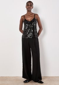 Schwarzes Pailletten-Camisole mit verstellbaren Spaghettiträgern, kombiniert mit luftigen schwarzen weiten Hosen und glänzenden schwarzen Schuhen.