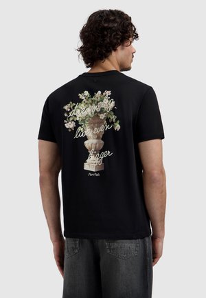 Man met krullend haar, gekleed in een zwart T-shirt met een grafische afbeelding van een bloemenvaas en de tekst "dream big live even bigger" op de achterkant, gecombineerd met een zwarte spijkerbroek.