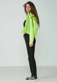 Neon-grüner, verkürzter Hoodie mit Reißverschluss und Kordelzügen, kombiniert mit schwarzen Hosen und schwarzen Sneakers. Glatte Texturen; lässiges Design.