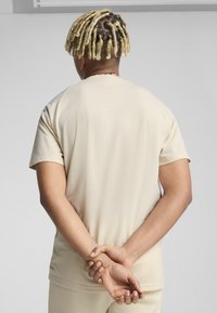 Beżowy t-shirt z krótkim rękawem i okrągłym dekoltem. Miękka tkanina o gładkiej strukturze. Nosi się z rękami złożonymi za plecami.