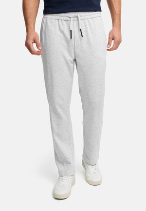 Mężczyzna w jasnoszarych spodniach dresowych z troczkiem i białych trampkach, stojący na jednolitym białym tle.