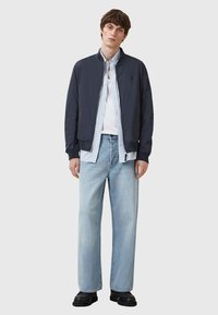 Marine bomberjakke over en lyseblå skjorte med knapper, parret med lyseblå vide jeans og svarte sko. Enkelt design, ingen synlige mønstre.
