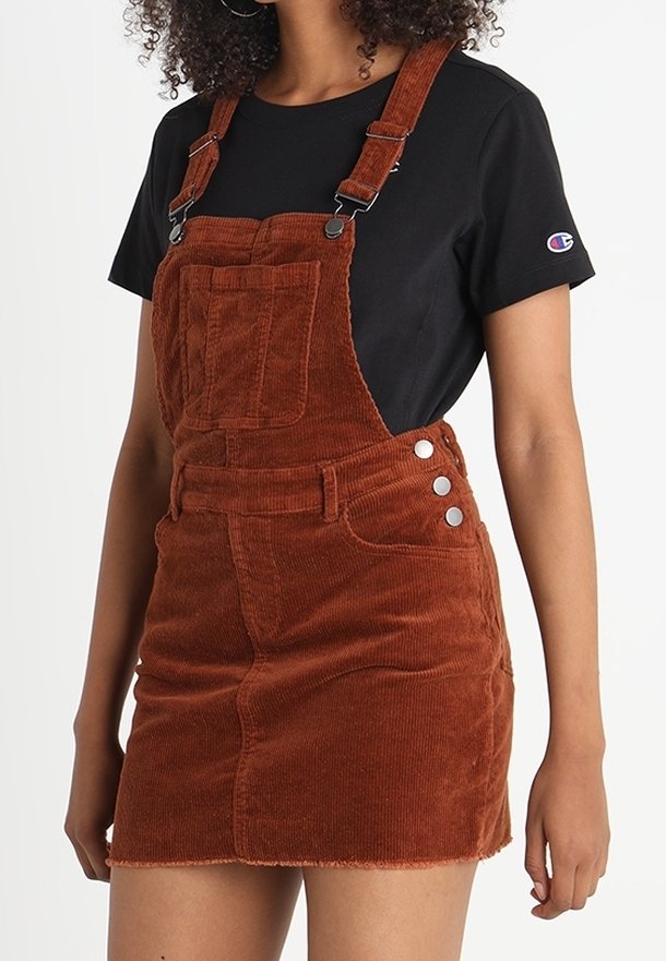Femme portant un t-shirt noir à manches courtes et une robe salopette en velours côtelé marron avec une poche avant et des boutons argentés sur le côté.