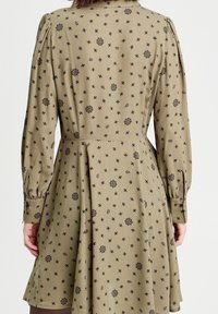 Femme portant une robe beige avec de longues manches bouffantes et des motifs floraux et de feuilles noirs, vue de dos.