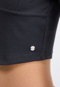 Black ribbed fabric with a fitted design, featuring a silver circular logo accent and contrasting stitching along the hem.