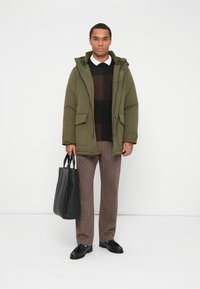 Férfi zöld kapucnis dzsekiben, fekete pulóverben, barna nadrágban és fekete cipőben, fekete táskával a kezében, fehér háttér előtt áll.