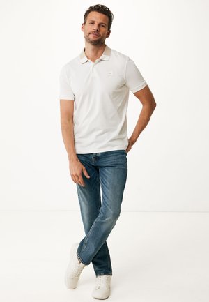 Uomo in piedi con le gambe incrociate, indossa una polo bianca, jeans blu e sneakers bianche su uno sfondo bianco uniforme.