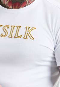 Vit bomullströja med guld broderad text "SILK" på bröstet. Rundad halsdesign och slät textur. Inga ytterligare mönster eller accenter.