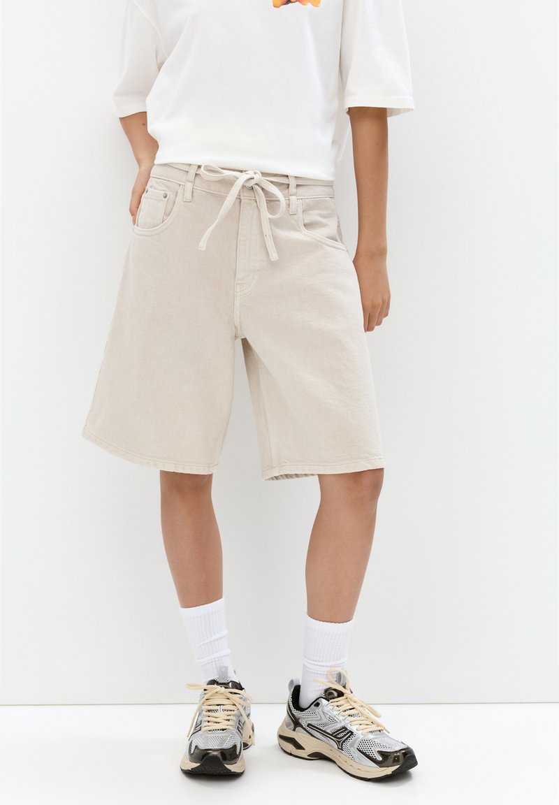 Beige denimshorts med avslappnad passform, bälte i midjan och sidofickor, kombinerat med en vit t-shirt och sportskor med svarta detaljer.