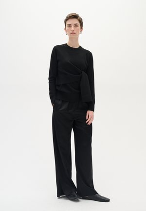 Černý svetr s dlouhým rukávem a zkrouceným designem v pase, spárovaný s volnými černými kalhotami a černými nazouvacími botami. Minimalistický styl.