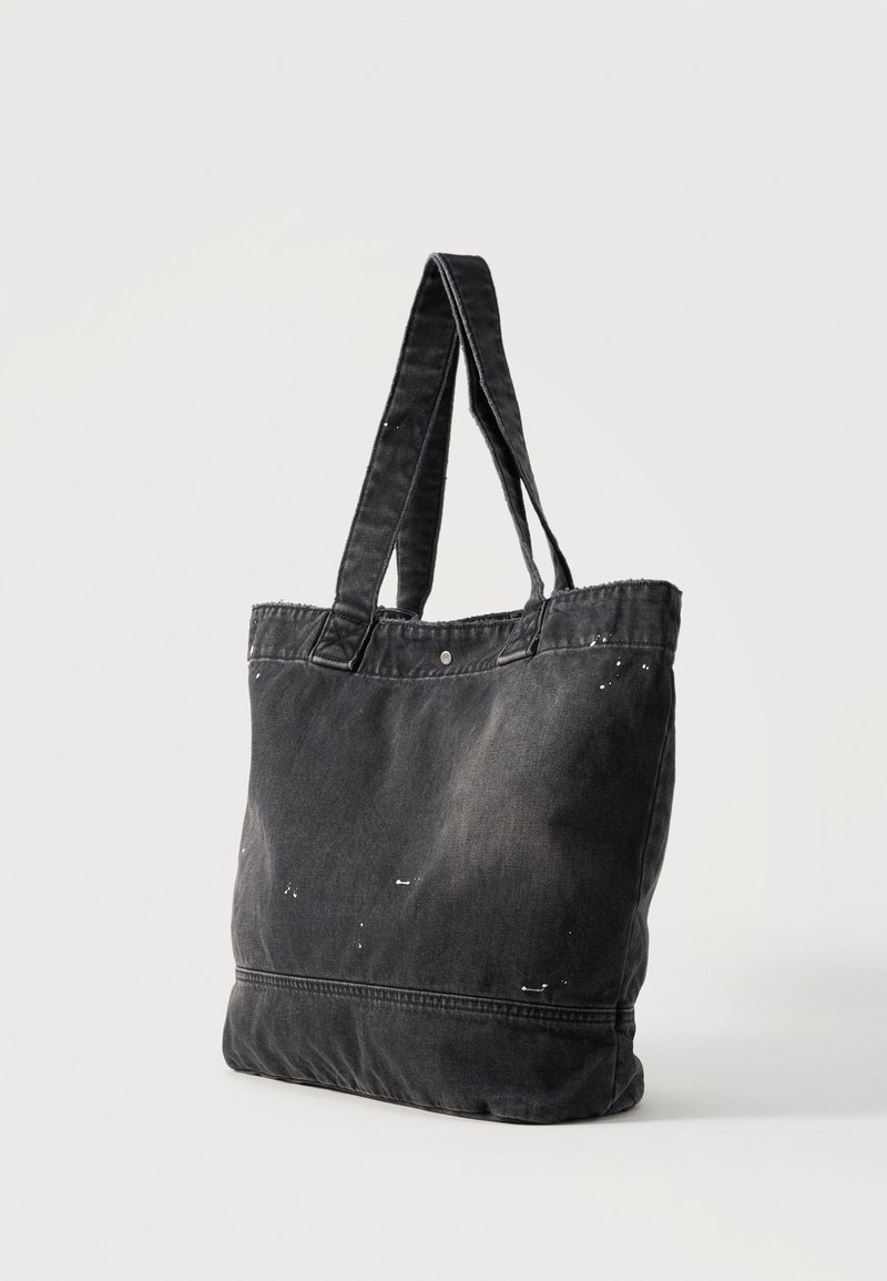 Schwarze Stofftasche mit zwei Schulterriemen, einem Druckknopfverschluss und dezenten weißen Farbspritzern auf der Oberfläche vor einfarbigem Hintergrund.