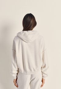 Felpa con cappuccio in pile bianca con vestibilità comoda, con cappuccio morbido leggermente arricciato e polsini a coste. Il materiale sembra spesso e confortevole.