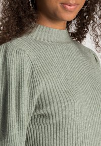 Vihreä ribbineulepaita, jossa on korkea kaulus ja teksturoitu kangas. Paita näyttää pystysuorat raidat ja istuvan siluetin.