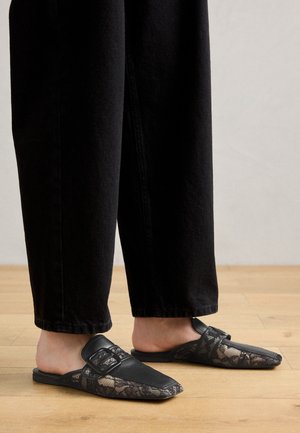 Mustad mustad nahast sussid, millel on pitsaktsendid, ümar varvas ja konksudetail. Kandmine laia musta püksiga. Puitpõranda taust.