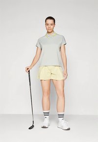 Jasnoszara koszulka polo z żółtym kołnierzykiem, zestawiona z żółtymi szortami. Czarny kij golfowy trzymany w jednej ręce, białe trampki z czarnymi paskami widoczne.