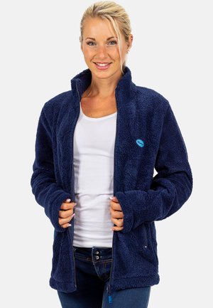 Frau lächelt, während sie eine marineblaue Fleecejacke über einem weißen Hemd und blauen Jeans trägt und die Reißverschlusskanten der Jacke hält.