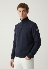 Marineblauer Reißverschluss-Pullover mit hohem Kragen, gerippten Bündchen und Saum, mit einem Logo-Patch am linken Ärmel. Das Material wirkt weich und strukturiert.