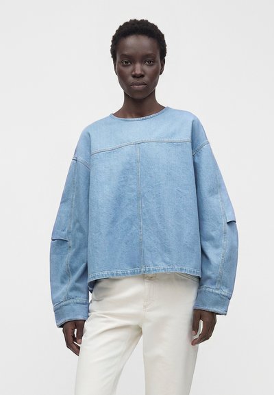 Femme aux cheveux courts et bouclés portant un sweat-shirt ample en denim bleu clair et un pantalon blanc, debout devant un fond uni.