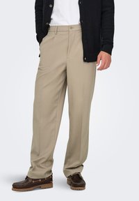 Pantalon beige sur mesure à coupe droite, associé à un cardigan noir et des bateaux marron. Tissu lisse avec des plis avant définis.