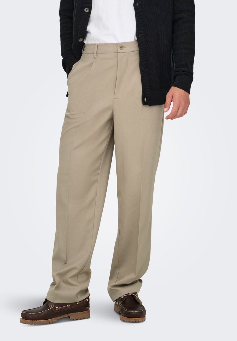 Pantalon beige sur mesure à coupe droite, associé à un cardigan noir et des bateaux marron. Tissu lisse avec des plis avant définis.