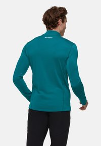 Langarm-Performance-Shirt in Türkis, aus elastischem Stoff mit Flachnähten und einem Logodetail auf der Rückseite. Glatte Textur, atletischer Schnitt.