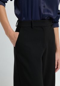Personne portant un pantalon noir sur mesure avec une main dans la poche et une blouse bleu marine à boutons avec les manches retroussées.