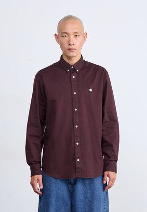 Bordeauxfarbene Button-up-Hemd mit weißen Knöpfen und einem kleinen Logo auf der Brust, kombiniert mit lockeren blauen Denim-Shorts. Glatte Stofftextur.