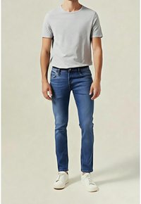 Man wearing light gray short-sleeve t-shirt, slim-fit blue jeans, and white sneakers against a plain light background.