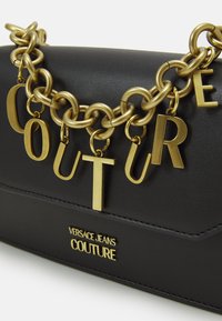 Černá kožená taška Versace Jeans Couture se silným zlatým řetězem a visícími zlatými písmeny, která tvoří slovo "COUTURE."