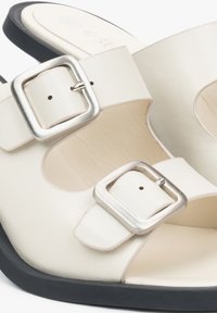 White leather sandals with two metallic buckles, open-toe design, and black soles. Smooth texture and minimalist style.