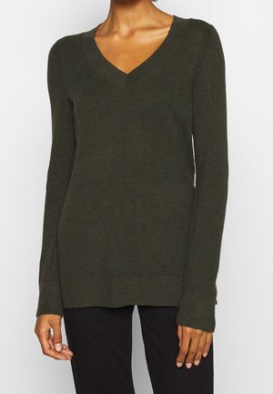 Femme portant un pull en maille vert foncé à manches longues avec un col en V et un motif texturé, associé à un pantalon noir, visible du cou aux hanches.