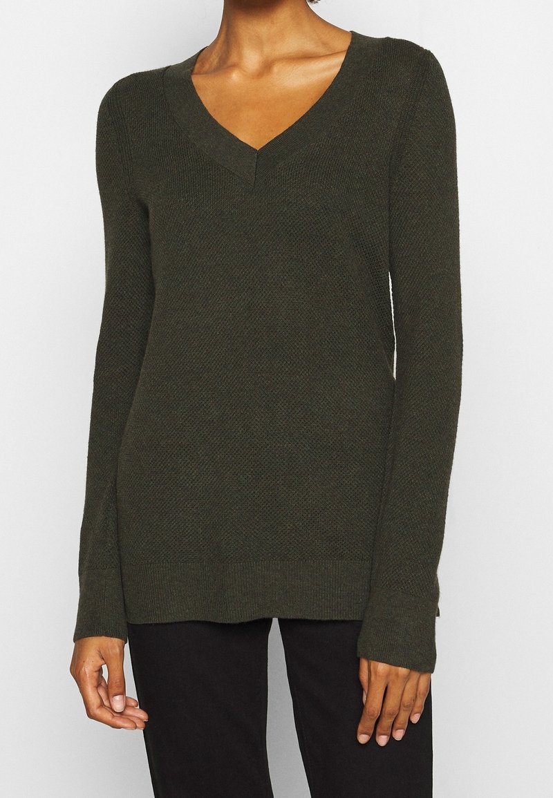Femme portant un pull en maille vert foncé à manches longues avec un col en V et un motif texturé, associé à un pantalon noir, visible du cou aux hanches.