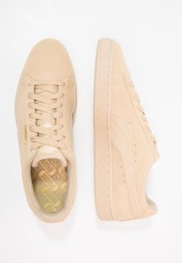 Sneakers in suède beige con lacci abbinati, suole in gomma e sole interne con motivo dorato, mostrati da viste dall'alto e di lato su uno sfondo bianco.