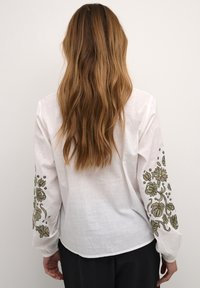 Camicia bianca a maniche lunghe con ricami floreali verdi sui polsini. Realizzata in tessuto leggero, presenta una vestibilità rilassata e una texture liscia.