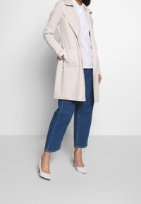 Beige mocka lång kappa med slag, sidofickor och avslappnad passform, tillsammans med blå denimbyxor och vita klackar.