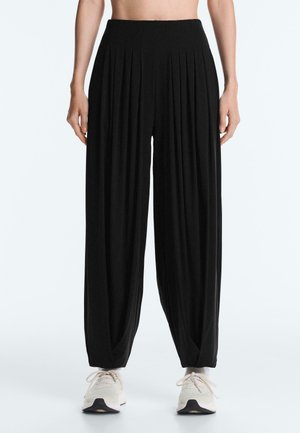 Čierne plisované nohavice s vysokým pásom a širokými nohavicami, nosené s bielymi teniskami, zobrazené od pása po nohy na jednoliatom pozadí.